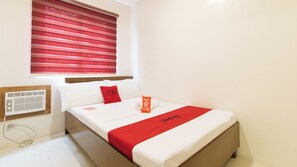 Una habitación de hotel con una cama, una ventana con persianas rojas, un almohadón blanco, un almohadón rojo, una colcha roja y una mesita de noche blanca con un radio.