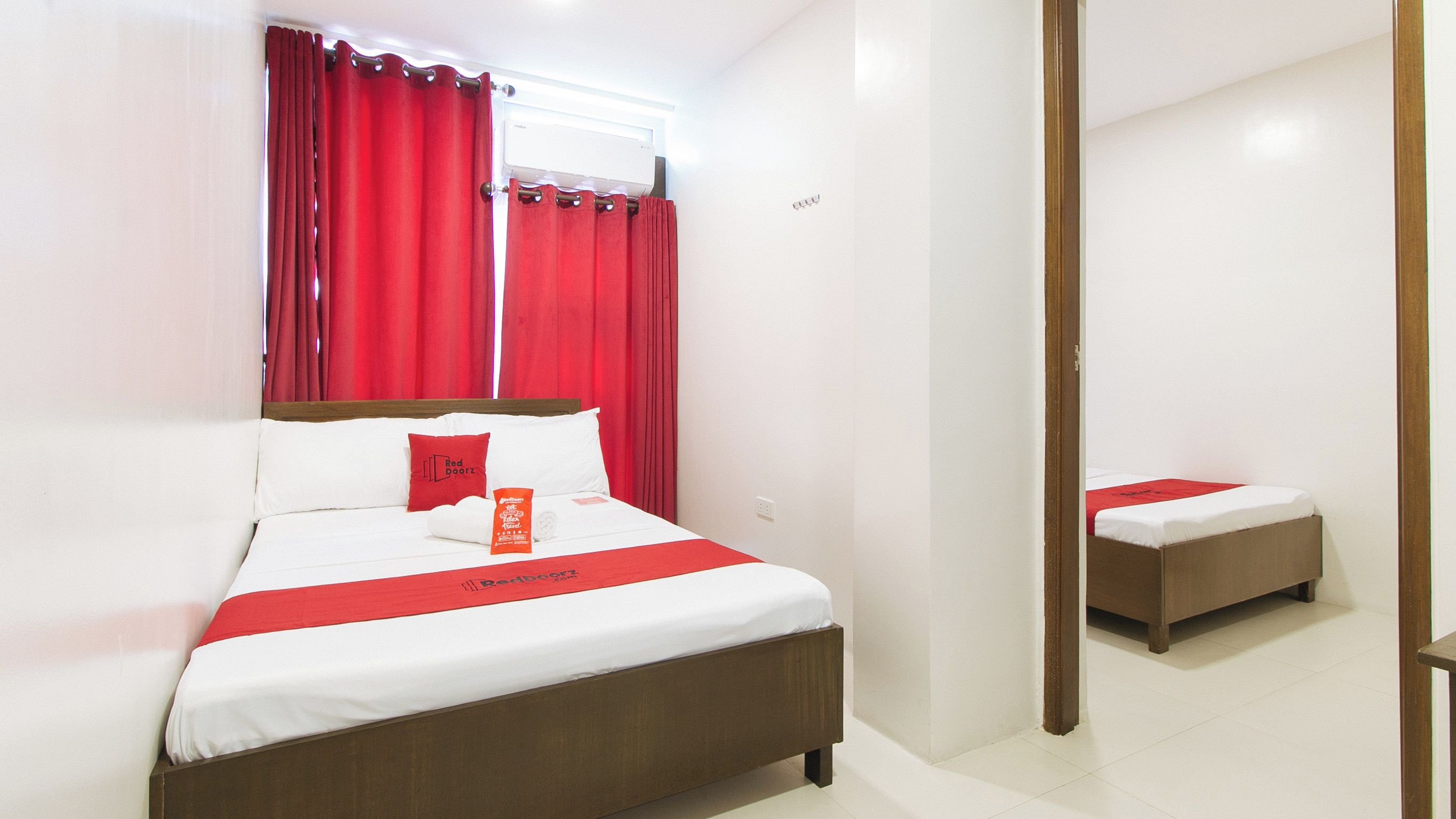 A hotel room with a large bed, a smaller bed, red curtains, a window with an air conditioner, and a wooden door.