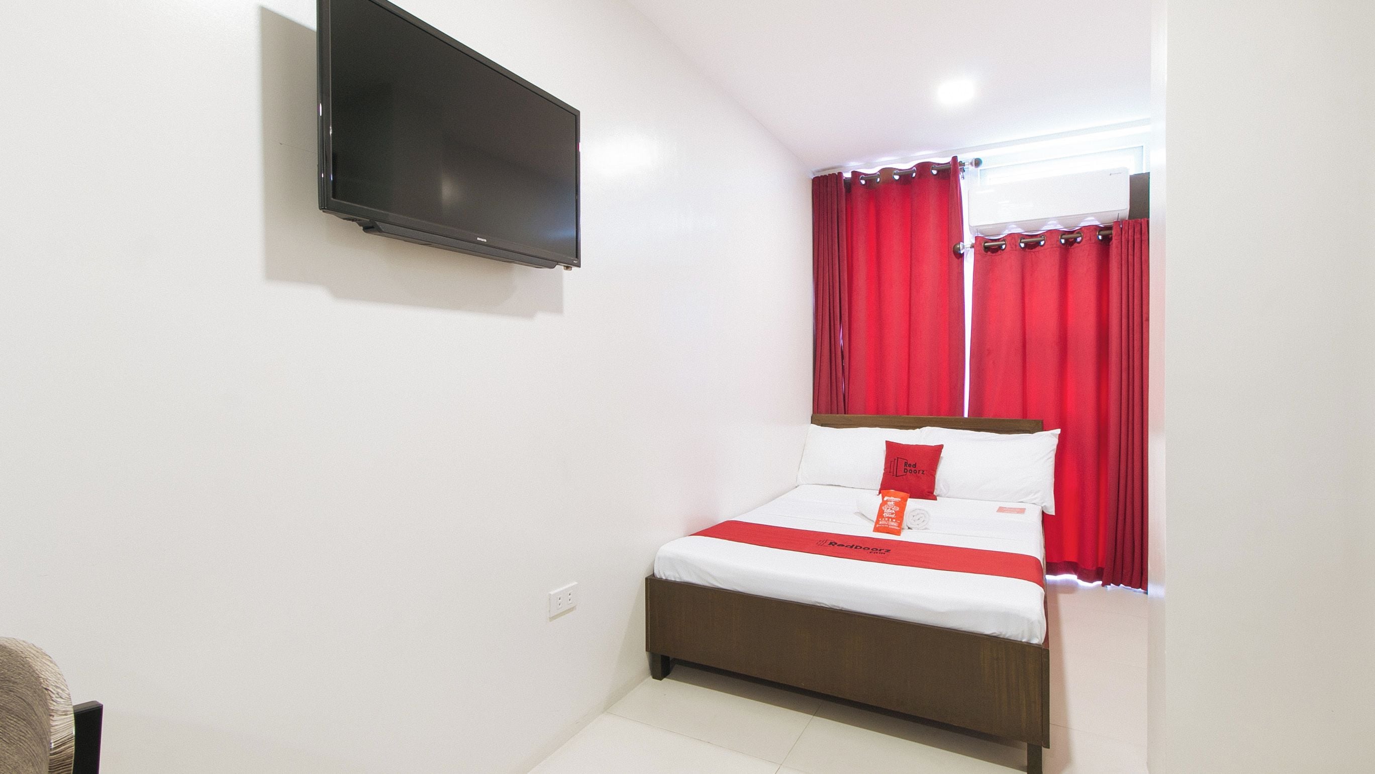 A hotel room with a bed, a television mounted on the wall, and red curtains.