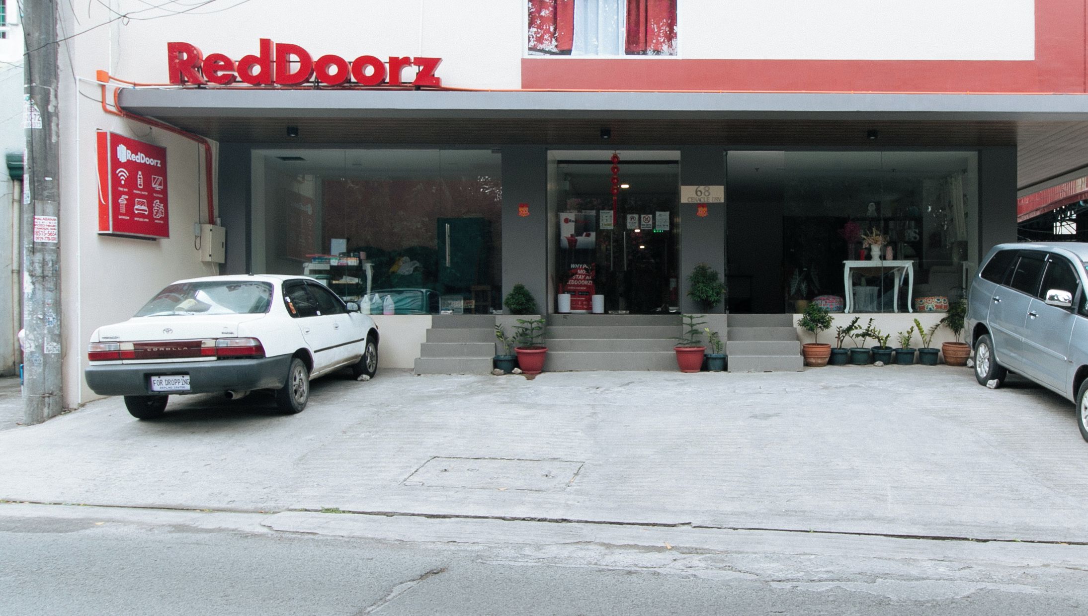 Un edificio RedDoorz con un acceso cubierto, dos coches aparcados y plantas en macetas al frente.