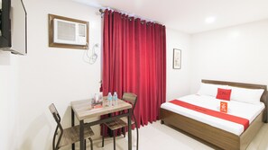 A hotel room with a bed, a table with chairs, a red curtain, and a mounted air conditioner.