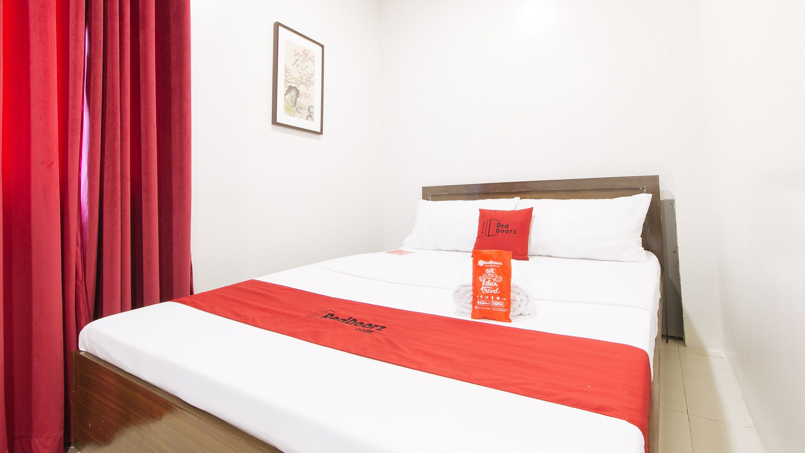 A hotel room with a neatly made bed, red curtains, and a framed picture on the wall.