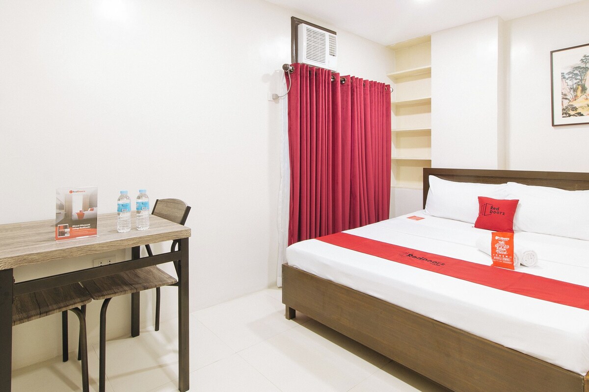 Una habitación de hotel con una cama, una mesa con botellas de agua, una cortina roja y un aire acondicionado de pared.