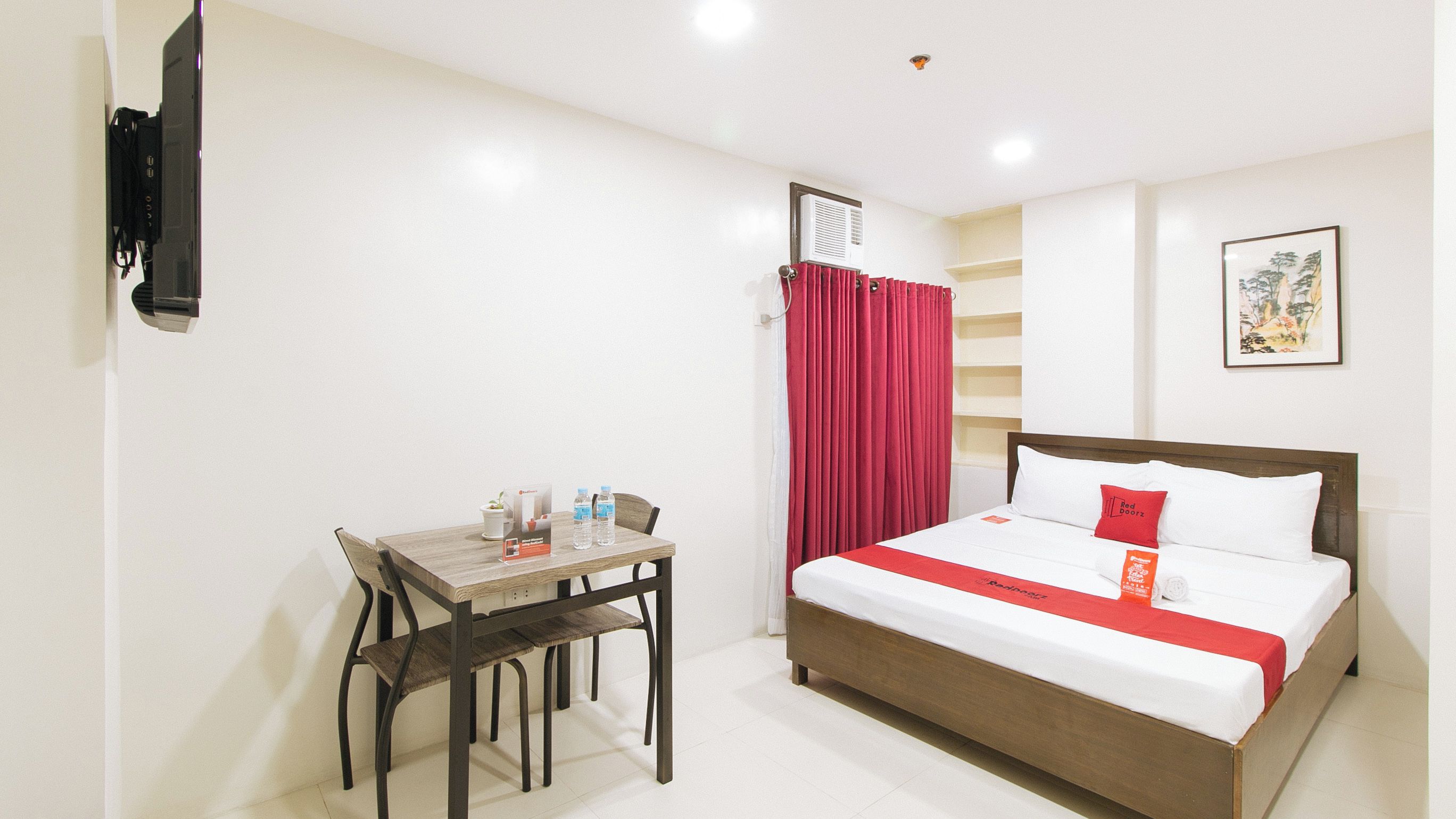 A hotel room with a bed, a desk with chairs, a TV, a red curtain, and a small plant.