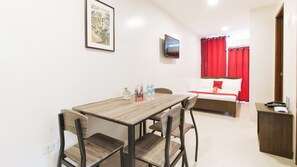 Habitación de hotel con una mesa de comedor de madera, dos sillas, una cama con cortinas rojas, un televisor en la pared y un escritorio pequeño con un teléfono.