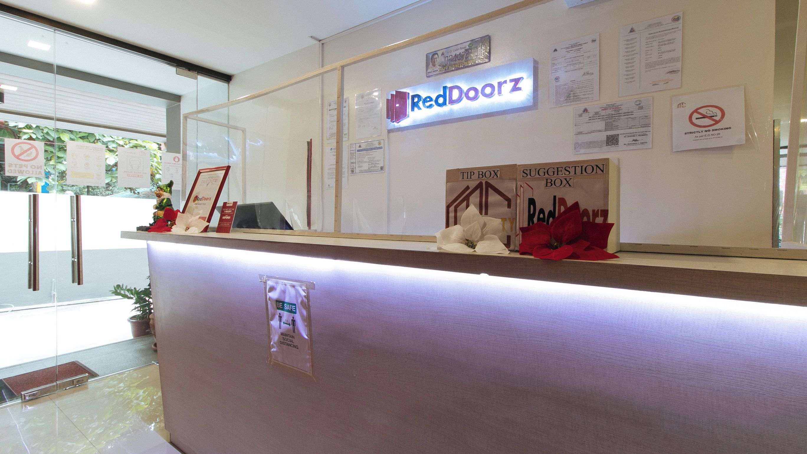 Un comptoir d’accueil avec une enseigne RedDoorz, un panneau « interdiction de fumer » et une affiche indiquant : « Restez prudents, respectez les distances de sécurité. »