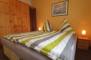 A neatly made bed with striped bedding, a wooden wardrobe, a nightstand with a lamp, and a framed picture on the wall. - TUI Haus Ahlbeck - Räuberkuhle 1 Vacation Homes (Koserow)
