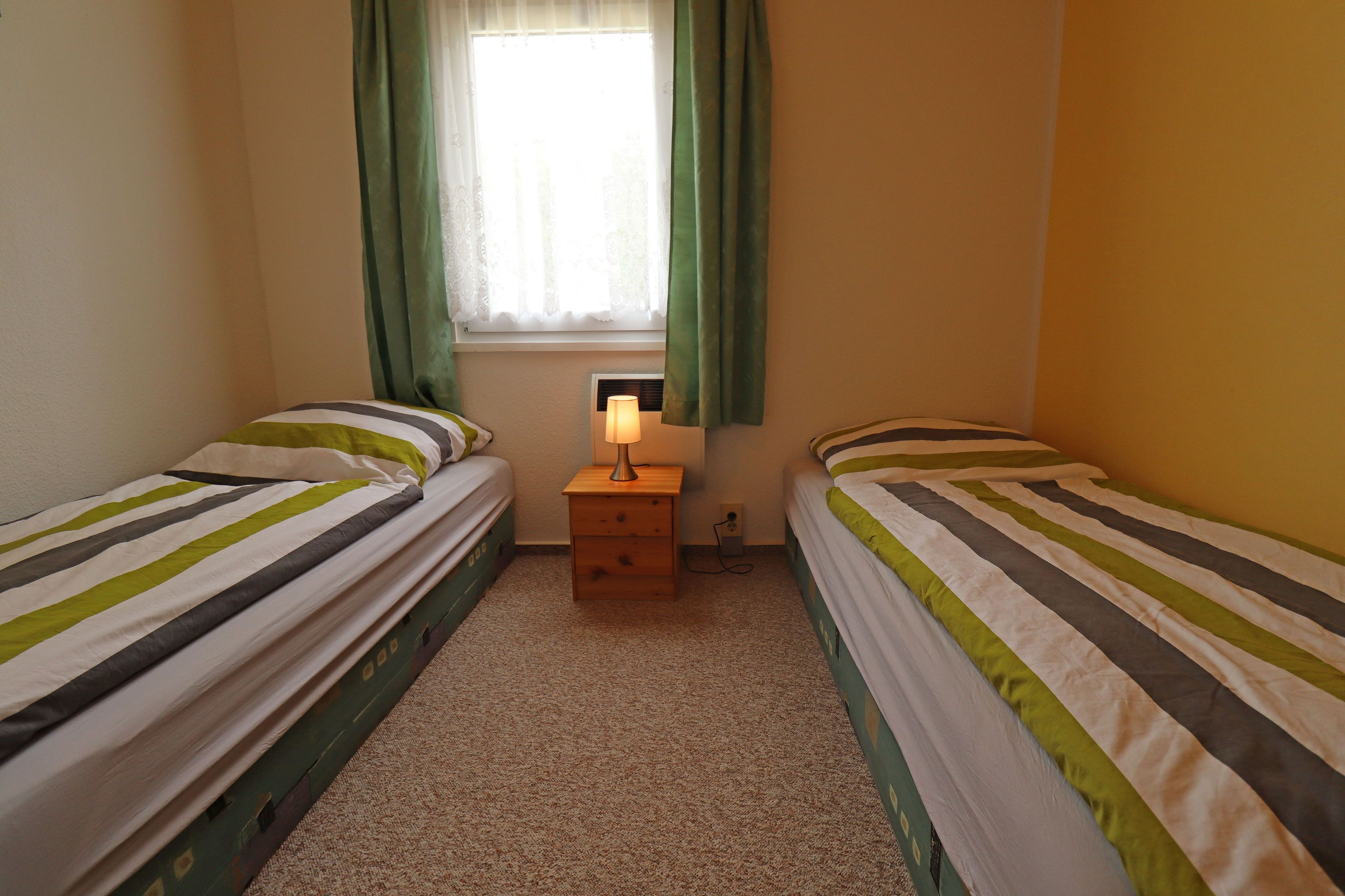 Une petite chambre agréable, dotée de deux lits simples, d’une table de chevet avec une lampe et d’une fenêtre avec des rideaux.