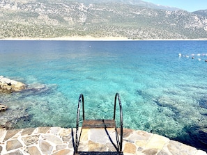 Ein Steingrund-Pool mit klarem, blauem Wasser, ein Holzdeck und Blick auf eine gebirgige Küstenlandschaft.