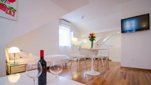 Een moderne, goed verlichte kamer met een houten vloer, een bed, een eettafel met stoelen, een aan de muur bevestigde televisie en een raam met gordijnen.
