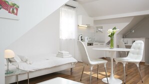 Een moderne, witte slaapkamer met een bed, nachtkastje en een zithoekje. Een eettafel met stoelen staat in het midden van de kamer, met een bloemenvaas erop.