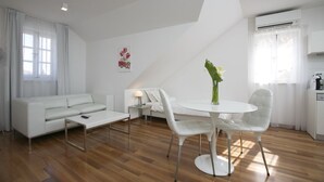 Een moderne woonkamer, ingericht in witte tinten, met een eethoek, houten vloer en een groot raam met gordijnen.