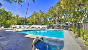 Una piscina con un pequeño jacuzzi, rodeada de palmeras y sillones de descanso.
