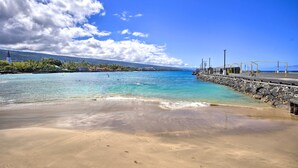 Una playa con agua turquesa cristalina, un rompeolas rocoso y una cadena montañosa distante bajo un cielo parcialmente nublado.