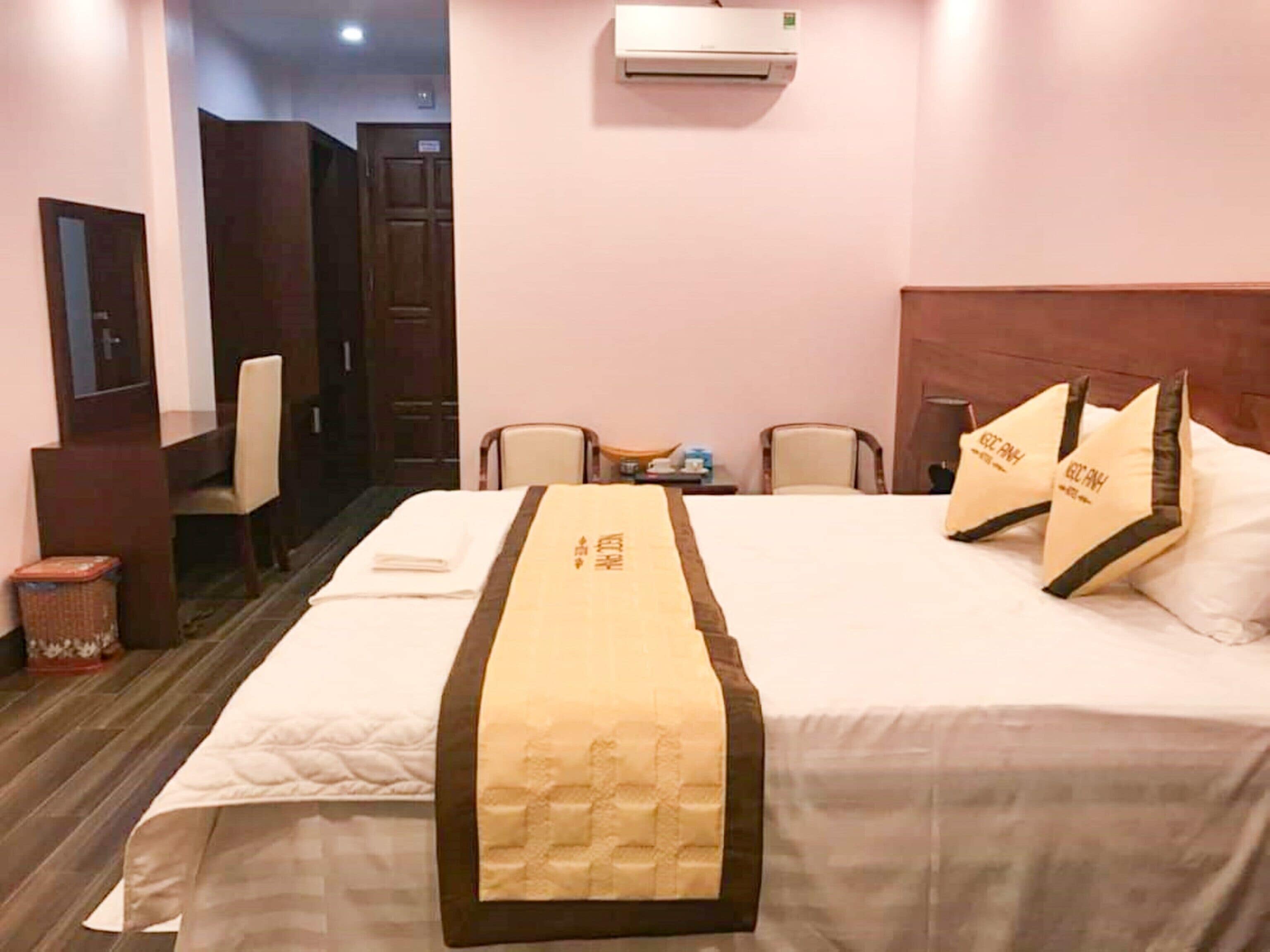 A hotel room with a neatly made bed, a bedside table with a lamp, a chair, and an air conditioning unit on the wall.