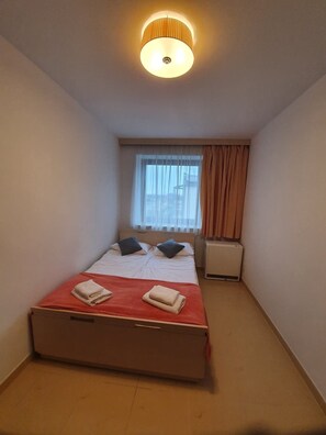 A small, well-lit bedroom with a single bed, a bedside table, a window with curtains, and a ceiling light. - LIDO Apartamenty (Sarbinowo)