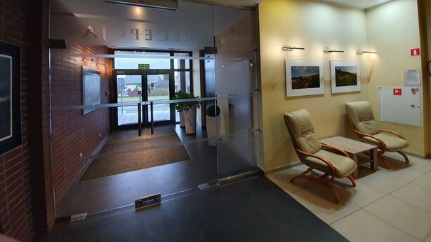 Zona de recepción con entrada por puerta de vidrio, mostrador de recepción, dos sillones acolchados y detector de incendios en la pared.