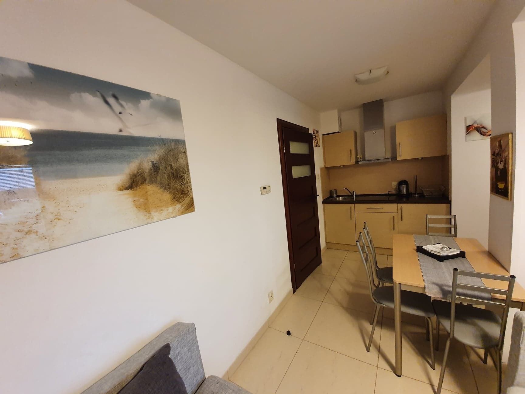 A compact kitchen with a dining area, a painting of a beach scene, and a door leading to another room.