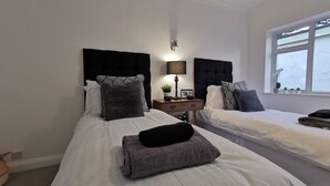A bedroom with two beds, a bedside table with a lamp, and a window with plants. - Beech Dreams - Stylish Place to Stay (Conwy)