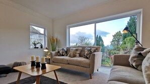 A modern living room with a sofa, a coffee table, and a lamp. There is a large window with a view of trees and a garden. - Beech Dreams - Stylish Place to Stay (Conwy)
