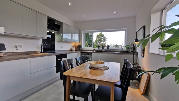 A modern kitchen with a dining area, featuring a wooden table and chairs, a built-in oven, and a window with a view of greenery. - Beech Dreams - Stylish Place to Stay (Conwy)