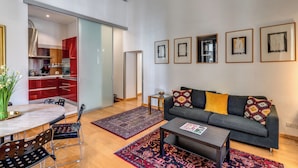 En moderne stue med en grå sofa, et sofabord og et spiseområde med bord og stole. Der er billeder i rammer på væggen, og et køkken kan ses gennem en åben dør.