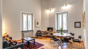En rummelig opholdsstue med et rundt spisebord, flere stole, en sofa, et fjernsyn på et stativ og en reol.