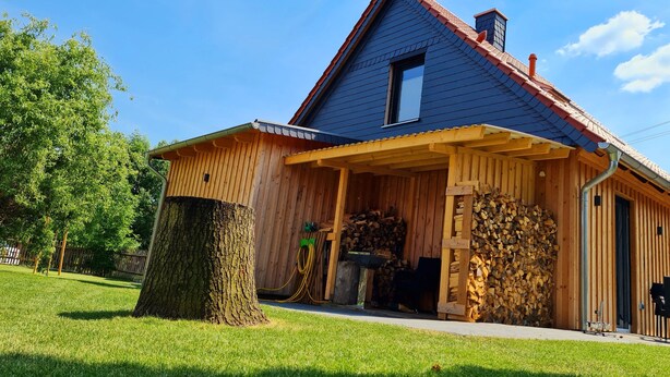A wooden house with a blue roof, a covered porch, and a stack of firewood outside.