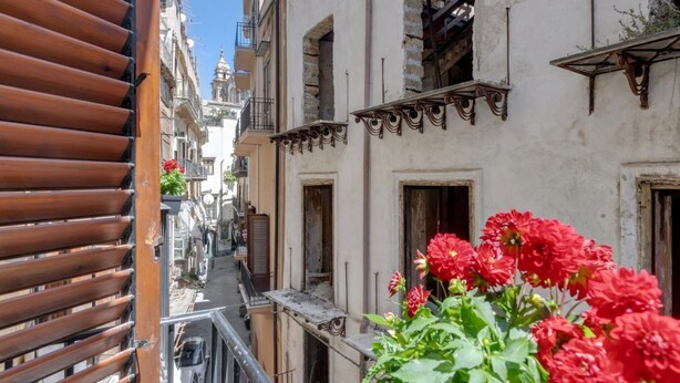 A narrow street with buildings on either side, balconies, and red flowers in the foreground. - GuestHost - Cozy 60 sqm apartment, for 3 people, with 3 balconies and located on the 2nd floor of a renovated building without lift. The property is located in the heart of the historic center (Traffic Restricted Area), inside Vucciria area, one of the c (Palermo)