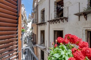 Una calle estrecha flanqueada por edificios, con balcones y flores rojas en primer plano.