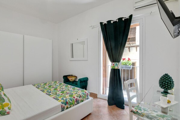Un dormitorio con cama, una silla, una mesita, una ventana con cortinas y un balcón con plantas.