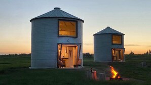 Two cylindrical yurts with open doors, one with a visible interior including a bed and a chair, and a small fire pit in the foreground. - Clark Farm Silos #1 - Panoramic Mountain Views (Kalispell)