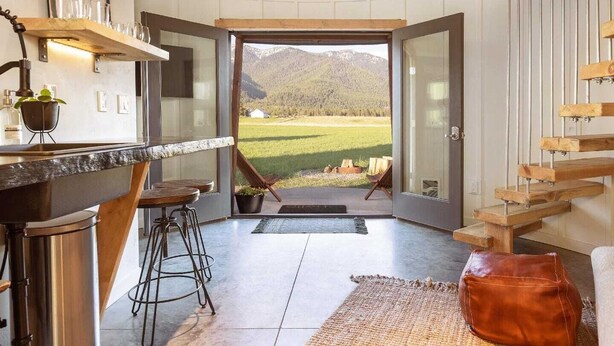 A modern kitchen with a bar area, a dining space with stools, and a view of a green landscape through an open door. - Clark Farm Silos #2 - Incredible  Mountain Views (Kalispell)