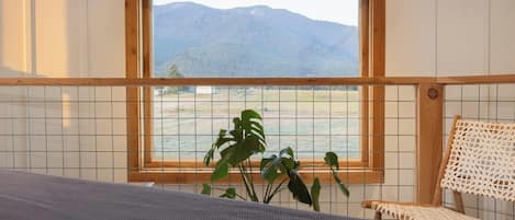 A room with a bed, a window with a view of mountains, a plant, and a woven chair.