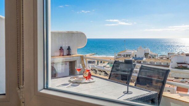 Een balkon met een tafel, twee glazen rode wijn en een fles, met uitzicht op de kustlijn met gebouwen en de zee.