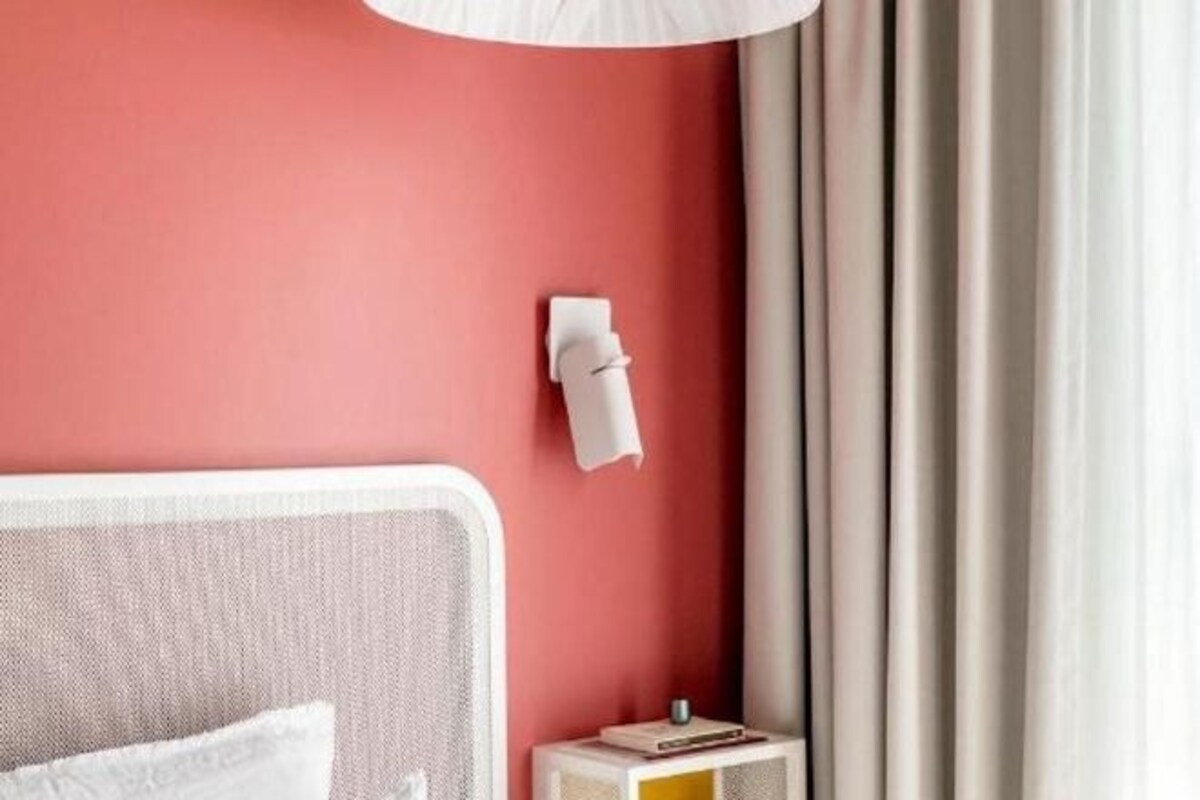 Une chambre avec un lit, une table de chevet sur laquelle se trouve un livre, une lampe fixée au mur et un rideau.