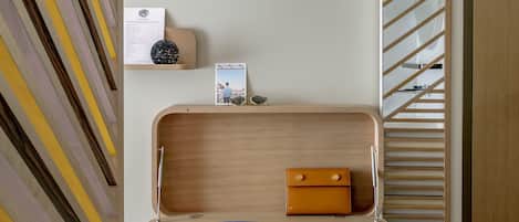 Una habitación moderna y minimalista con un muro de diseño geométrico, un escritorio de madera con una computadora portátil, un cuaderno y un bolígrafo, y una silla blanca.