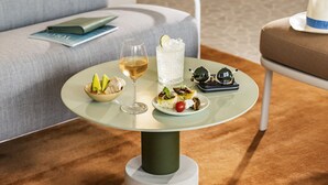 Una mesa de vidrio con un frutero, una copa de vino y un plato de comida.