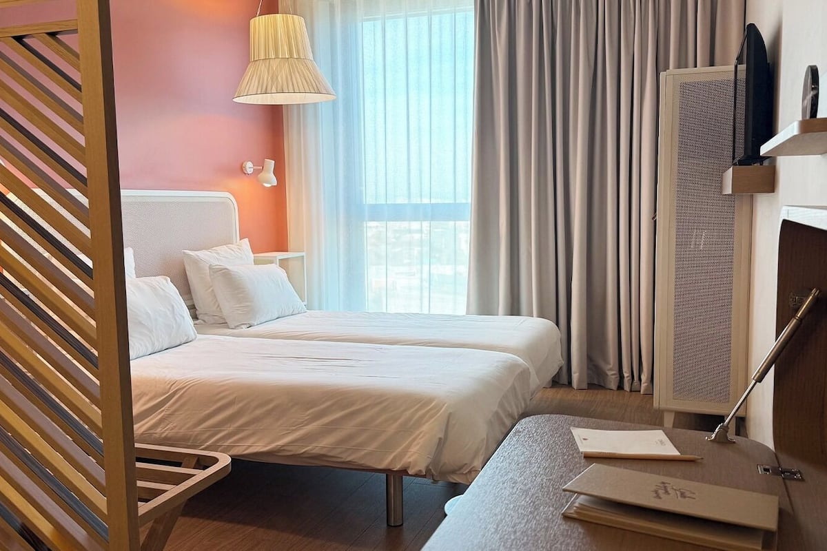 A hotel room with two beds, a desk, a TV, and a window with curtains.