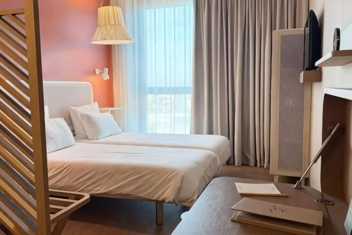 A hotel room with two beds, a desk, a lamp, a TV, and a window with curtains.