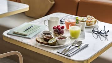 A breakfast table with a laptop, notebooks, a mug, bowls of food, a glass of orange juice, and a pair of glasses.