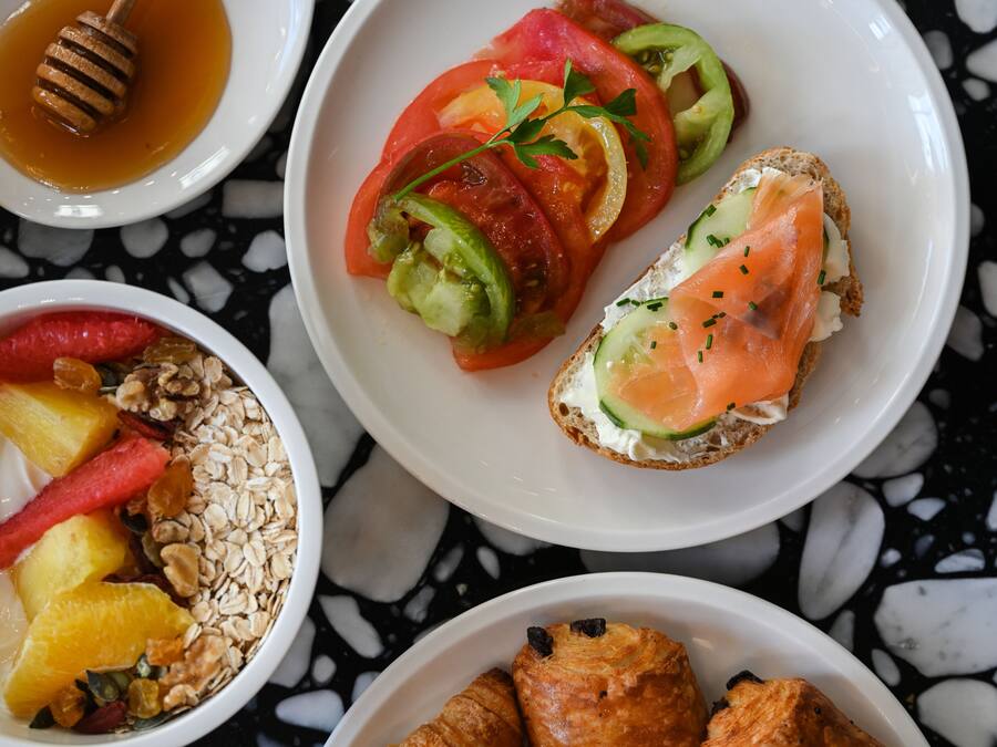 A breakfast setting with a variety of food items including a plate with sliced tomatoes, cucumbers, and a slice of smoked salmon on toast, a bowl of yogurt with fruits and granola, and a plate with croissants.