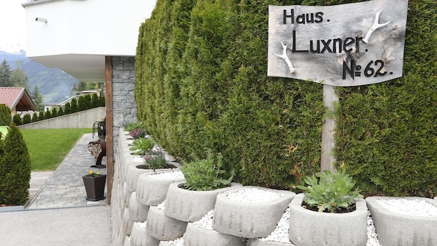 Une plaque portant l’inscription « Haus. Luxner. N° 62 » est fixée à la façade d’une maison dont le mur est en pierre et qui est agrémentée de plantes en pot.