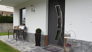 Une entrée moderne avec une porte sombre, une pancarte de bienvenue, un panier en osier rempli de fleurs et une petite plante en pot.
