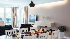 A modern dining area with a white table set for four, a black hanging light fixture, and a flat-screen TV mounted on a wooden shelf.