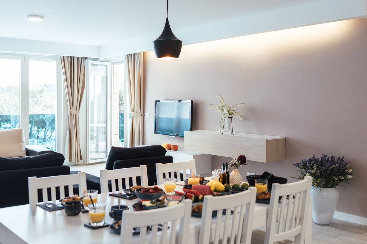 A modern dining area with a white table set for four, a black hanging light fixture, and a flat-screen TV mounted on a wooden shelf.