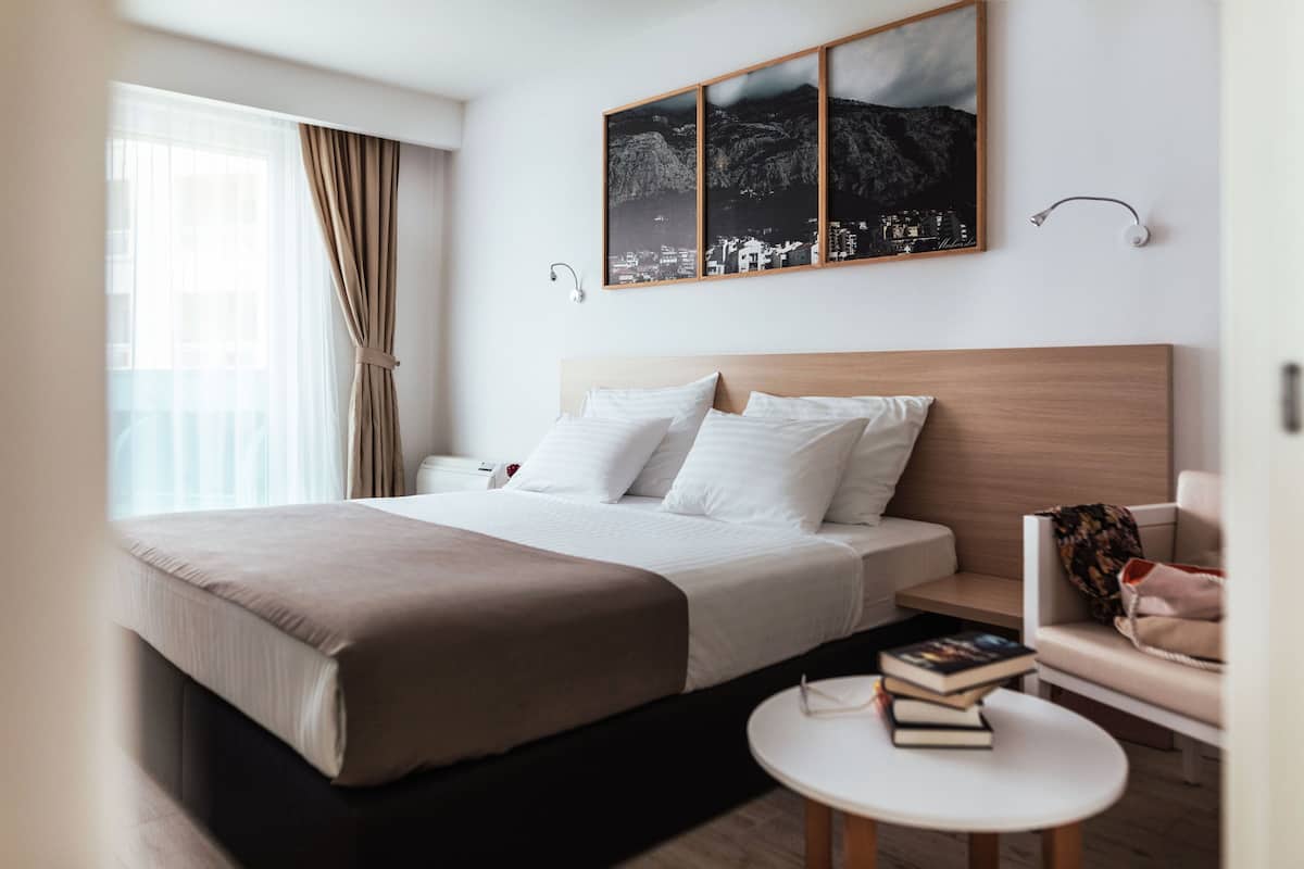A modern hotel room with a large bed, a bedside table with books, a sofa, and a cityscape artwork on the wall.