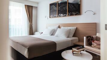 A modern hotel room with a large bed, a bedside table with books, a sofa, and a cityscape artwork on the wall.