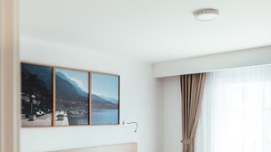A modern hotel room with a large bed, a headboard with a cityscape artwork, a window with sheer curtains, and a light wooden floor.