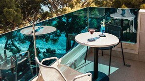 A glass-enclosed balcony with a table and chairs, overlooking a scenic view of trees and the sea.